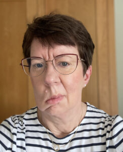 Karen is wearing a white and black striped top and glasses. She has brown, short hair and the photo shows her one sided facial palsy. 